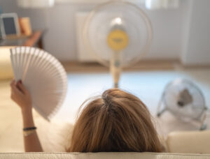 Menopausal woman sitting on a sofa with several fans giving her air and a fan in her hand.