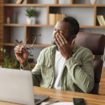 Tired sad mature black male takes off his glasses, rubbing eyes, suffering from overwork and overwork, discomfort long hours of sedentary in home office interior. Work, business and health problems