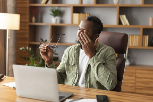 Tired sad mature black male takes off his glasses, rubbing eyes, suffering from overwork and overwork, discomfort long hours of sedentary in home office interior. Work, business and health problems